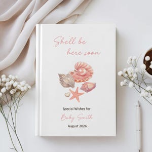 Peut inclure: Livre d'or de baby shower blanc avec le texte "She'll be here soon" et "Special Wishes for Baby Smith August 2026". La couverture présente des illustrations de coquillages roses et pêche, avec une étoile de mer et un cœur.