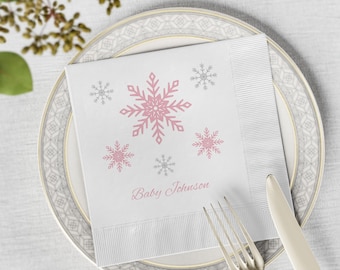 May include: A white square paper napkin featuring a pattern of pink and gray snowflakes and the text Baby Johnson printed in pink script. The napkin is placed on a white plate with a gray geometric border, accompanied by a silver fork and knife.