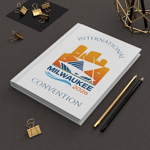 Op de afbeelding: Een wit hardcover notitieboek met de tekst "INTERNATIONAL MILWAUKEE CONVENTION 2026" in blauw en oranje. De omslag toont een gestileerde skyline en een golfontwerp. Gouden en zwarte potloden en paperclips liggen in de buurt.