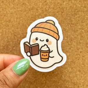 May include: A white ghost sticker wearing a brown beanie, reading a book, and holding a pumpkin spice latte. The sticker has a cute, cartoonish design with a fall theme. Other stickers are visible in the background.