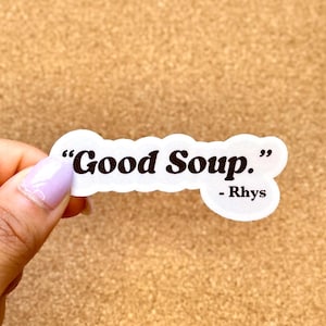 May include: A white sticker with the text "Good Soup." in black, with the name "- Rhys" below. The sticker is held by a hand with a light purple nail polish, set against a brown corkboard background.