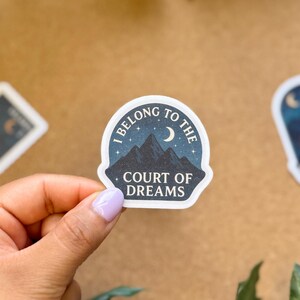 May include: A sticker with the text "I BELONG TO THE COURT OF DREAMS" in a circular design with a mountain and moon illustration. The sticker is held by a hand with a light purple nail polish. Other stickers are in the background.