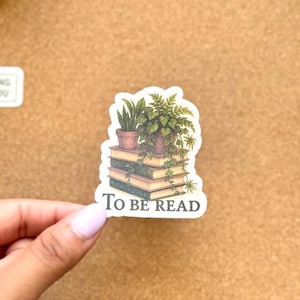 May include: A sticker with a stack of books and potted plants, with the text "TO BE READ". The sticker is held by a hand with a light pink nail polish. Other stickers with plants are also visible.