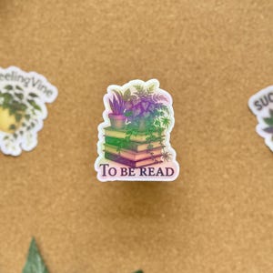 May include: A sticker featuring a stack of books with a small potted plant on top. The sticker has the words "TO BE READ" in white text. The books and plants have a gradient of green, purple, and pink.