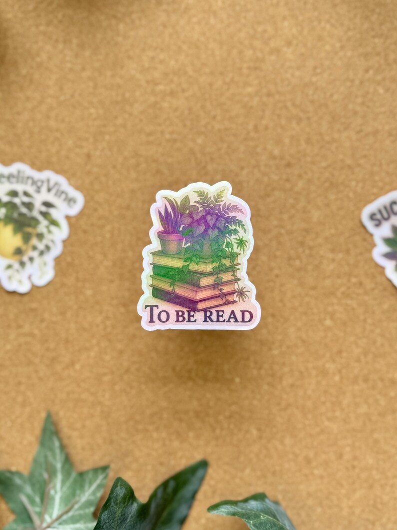 May include: A sticker featuring a stack of books with a small potted plant on top. The sticker has the words "TO BE READ" in white text. The books and plants have a gradient of green, purple, and pink.