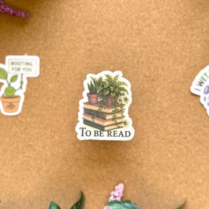 May include: A sticker featuring a stack of books with a potted plant on top. The sticker has the text "TO BE READ" at the bottom. The sticker is surrounded by green leaves and other stickers.