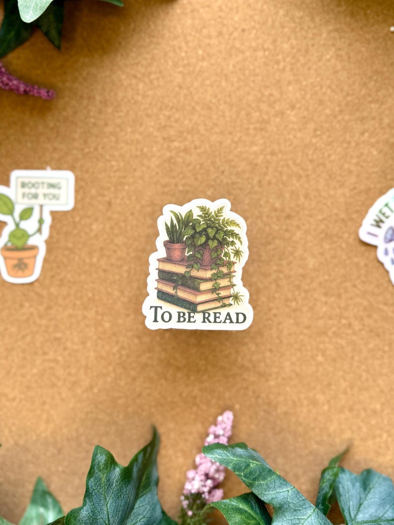 May include: A sticker featuring a stack of books with a potted plant on top. The sticker has the text "TO BE READ" at the bottom. The sticker is surrounded by green leaves and other stickers.