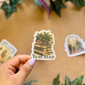 May include: A sticker featuring a stack of books with potted plants on top. The sticker has the words "TO BE READ" in white text. The sticker is held by a hand with a light purple nail polish.