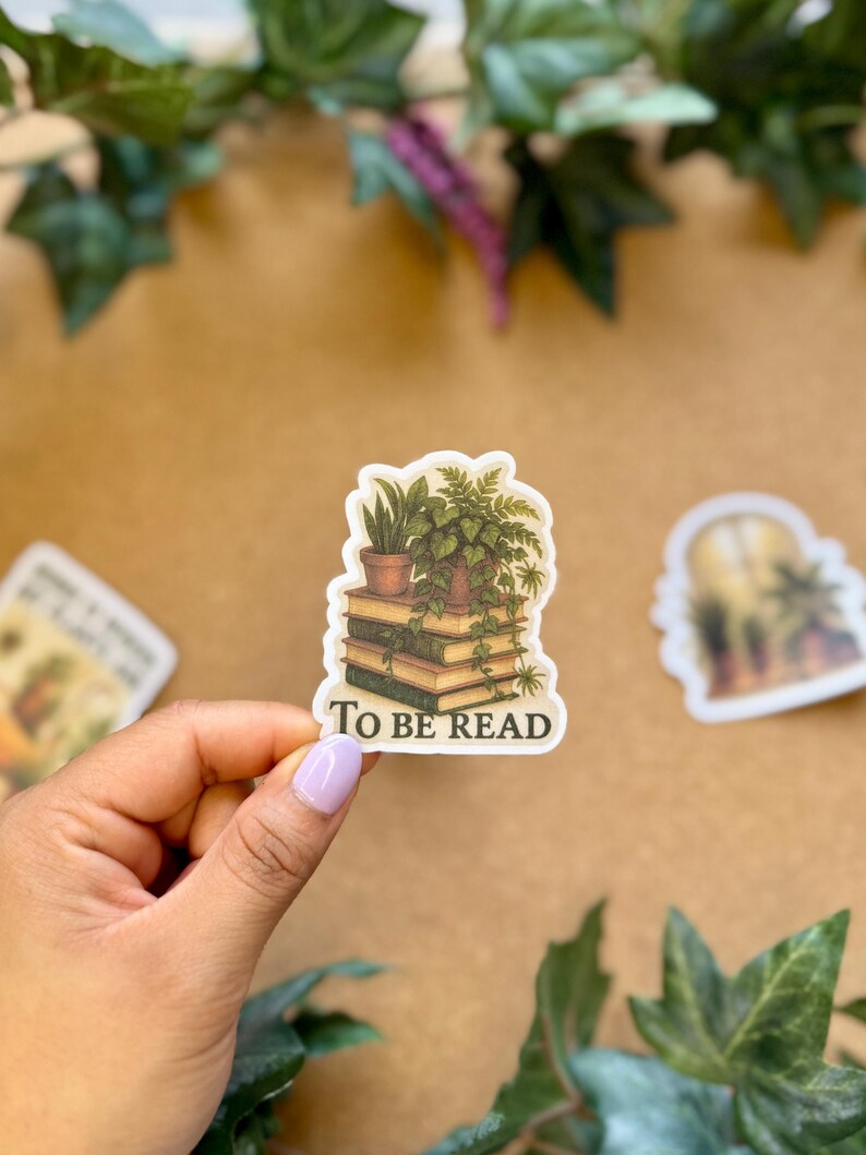 May include: A sticker featuring a stack of books with potted plants on top. The sticker has the words "TO BE READ" in white text. The sticker is held by a hand with a light purple nail polish.