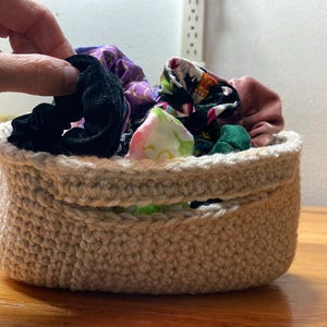 May include: A small, beige crocheted basket filled with colorful scrunchies. The scrunchies are various colors and patterns, including black velvet, floral prints, and solid colors. The basket is sitting on a wooden surface.