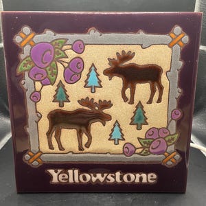 May include: A square ceramic tile with a purple border and a tan center. The tile features two brown moose, green and blue pine trees, and purple berries. The word "Yellowstone" is printed in white.