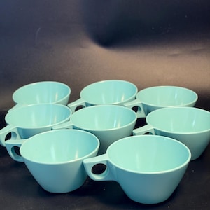 May include: A set of eight light turquoise cups arranged in a group. Each cup features a smooth, solid-colored finish, a wide circular rim, and a distinct geometric handle with a circular cutout, providing a uniform look across the entire collection.