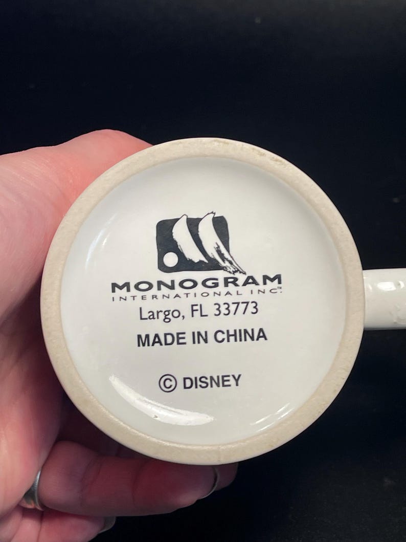 May include: White ceramic mug base with the text "MONOGRAM INTERNATIONAL INC." and "Largo, FL 33773". The text "MADE IN CHINA" and the Disney copyright symbol are also visible.