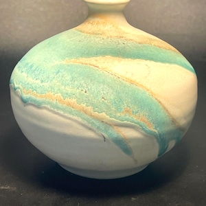 May include: A ceramic vase with a rounded body and a narrow neck. The vase features a white base, with turquoise and tan horizontal stripes. The top and neck are a light tan color. The vase has a smooth, glossy finish.