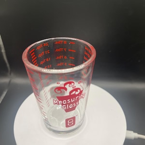 May include: A clear glass measuring cup with red measurement markings and the words "Measuring Glass" in red. The cup has a decorative design and the letter "S" in a red square. The cup is on a white surface.