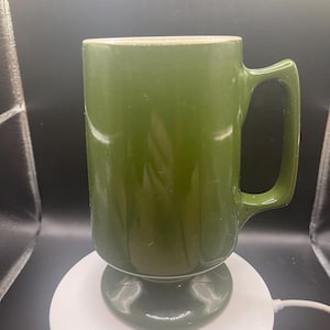 May include: A green ceramic mug with a pedestal base and a large handle. The mug has a cylindrical shape and a light-colored rim. The mug is approximately 6 inches tall.