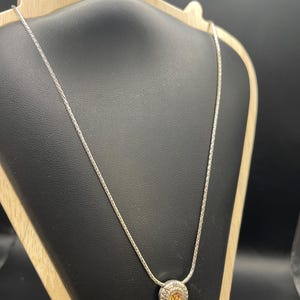 Op de afbeelding: Een zilveren ketting met een ronde hanger met een oranje edelsteen. De hanger is omgeven door een gedetailleerd, gestructureerd ontwerp. De ketting wordt gepresenteerd op een zwarte sieradenstandaard tegen een donkere achtergrond.