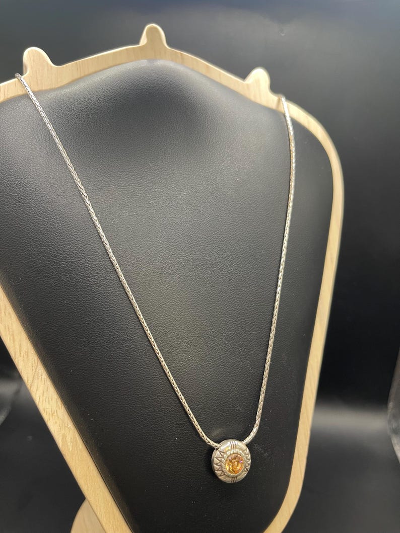 Op de afbeelding: Een zilveren ketting met een ronde hanger met een oranje edelsteen. De hanger is omgeven door een gedetailleerd, gestructureerd ontwerp. De ketting wordt gepresenteerd op een zwarte sieradenstandaard tegen een donkere achtergrond.