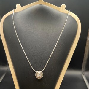 Op de afbeelding: Een zilveren ketting met een ronde hanger met een centrale heldere steen omgeven door een gedetailleerd, cirkelvormig ontwerp. De hanger hangt aan een delicate ketting, gepresenteerd op een zwarte sieradenstandaard.