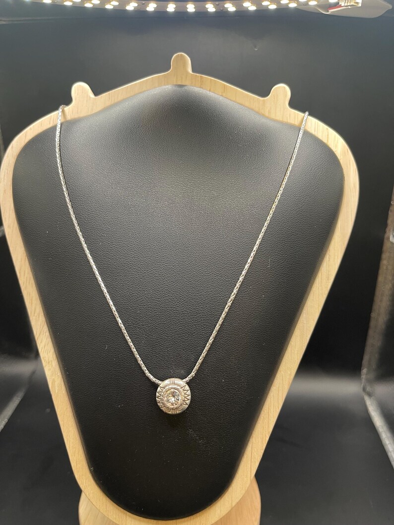 Op de afbeelding: Een zilveren ketting met een ronde hanger met een centrale heldere steen omgeven door een gedetailleerd, cirkelvormig ontwerp. De hanger hangt aan een delicate ketting, gepresenteerd op een zwarte sieradenstandaard.
