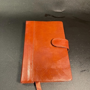 May include: A rectangular notebook with a smooth, reddish-brown cover featuring a vertical stitched seam along the left side. The notebook includes a matching reddish-brown strap closure on the right edge and a thin ribbon bookmark extending from the bottom.