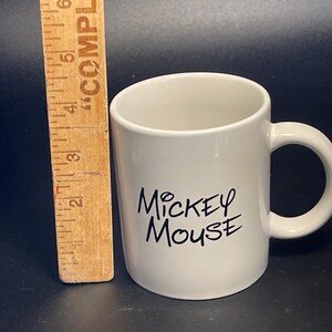 May include: A white ceramic mug with the words "Mickey Mouse" in black script. The mug has a curved handle and is approximately 3 inches tall. A wooden ruler is next to the mug.