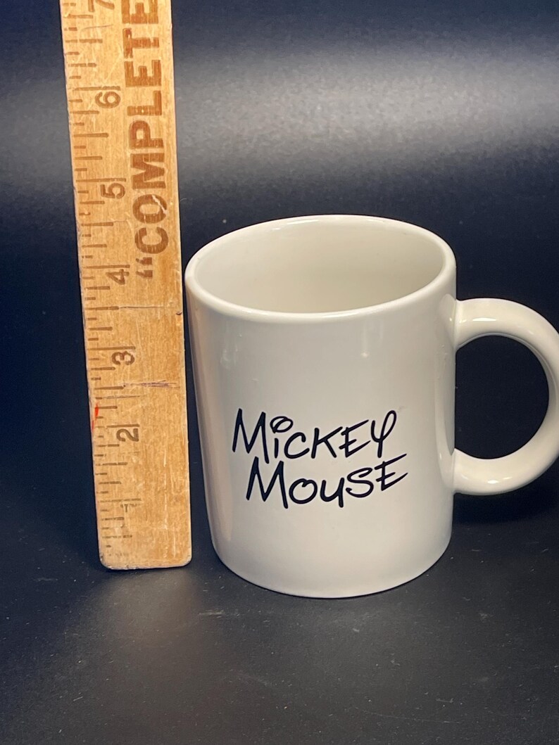 May include: A white ceramic mug with the words "Mickey Mouse" in black script. The mug has a curved handle and is approximately 3 inches tall. A wooden ruler is next to the mug.