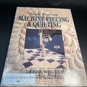 May include: A softcover instructional book titled Teach Yourself Machine Piecing & Quilting by Debra Wagner. The cover features a photograph of a sewing machine needle stitching a blue and white checkered quilt, with a white library sticker.