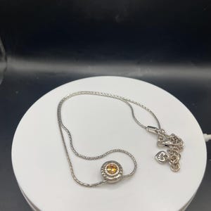 Op de afbeelding: Een zilverkleurige ketting met een kleine, ronde hanger met een gele edelsteen. De hanger is omgeven door een decoratieve rand. De fijne ketting heeft een hartvormige bedel bij de sluiting.