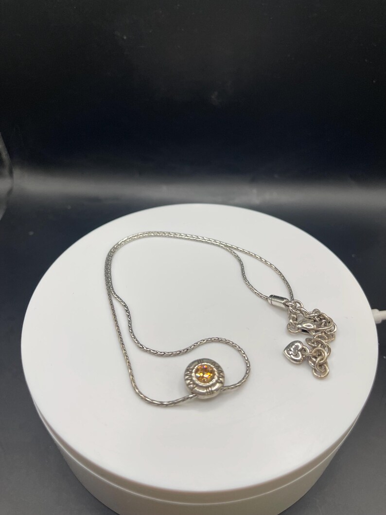 Op de afbeelding: Een zilverkleurige ketting met een kleine, ronde hanger met een gele edelsteen. De hanger is omgeven door een decoratieve rand. De fijne ketting heeft een hartvormige bedel bij de sluiting.