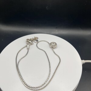 Op de afbeelding: Een zilveren ketting met een fijne ketting en een ronde hanger. De hanger heeft een textuurpatroon met een kleine, heldere steen in het midden. De ketting wordt gepresenteerd op een wit oppervlak tegen een zwarte achtergrond.
