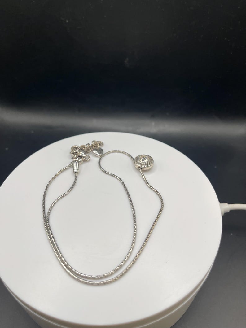 Op de afbeelding: Een zilveren ketting met een fijne ketting en een ronde hanger. De hanger heeft een textuurpatroon met een kleine, heldere steen in het midden. De ketting wordt gepresenteerd op een wit oppervlak tegen een zwarte achtergrond.