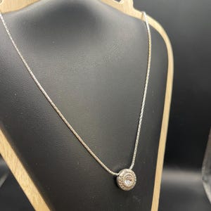 Op de afbeelding: Een zilveren ketting met een ronde hanger met een heldere edelsteen. De hanger heeft een decoratief, cirkelvormig ontwerp. De ketting wordt gepresenteerd op een zwarte sieradenstandaard met een houten voet.