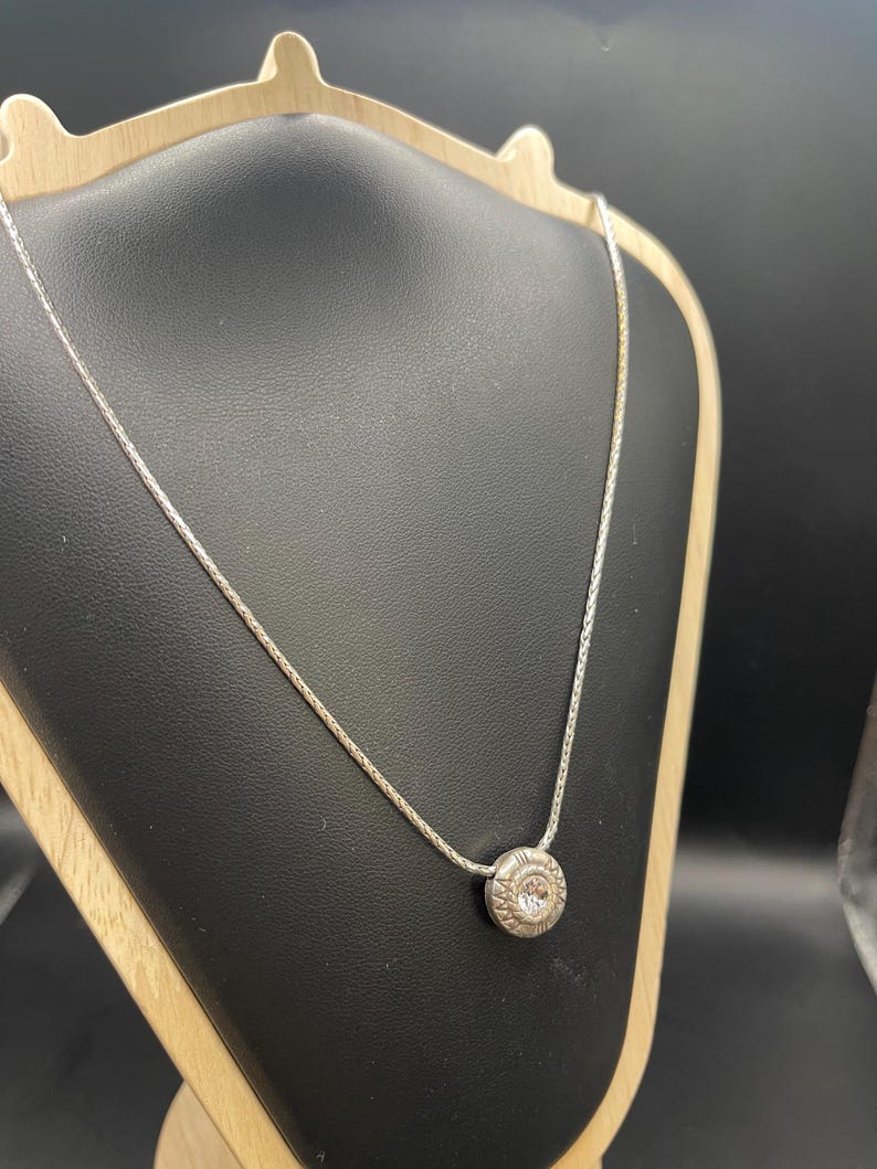 Op de afbeelding: Een zilveren ketting met een ronde hanger met een heldere edelsteen. De hanger heeft een decoratief, cirkelvormig ontwerp. De ketting wordt gepresenteerd op een zwarte sieradenstandaard met een houten voet.