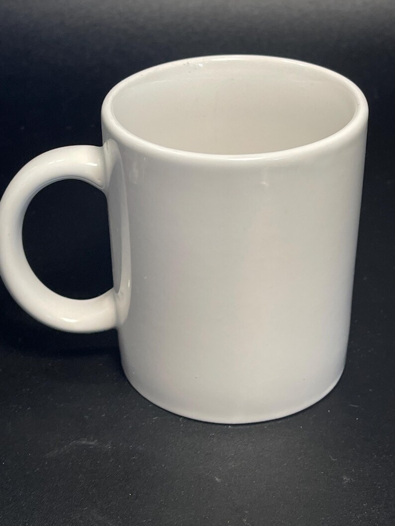 May include: A white ceramic mug with a rounded handle. The mug is cylindrical and has a smooth, glossy finish. The mug is empty and ready to be filled with a beverage.