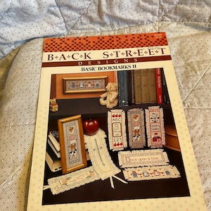 May include: A book titled "BACK STREET DESIGNS BASIC BOOKMARKS II" with various cross-stitch bookmark designs. The designs include framed pictures, bookmarks with bears, cows, and the text "ABC 123". The cover has an orange and white pattern.