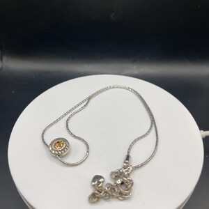 Op de afbeelding: Een zilveren ketting met twee verschillende hangers. E&eacute;n hanger is een cirkelvormig ontwerp met een gele edelsteen in het midden. De andere hanger is een cluster van zilveren vormen. De ketting wordt gepresenteerd op een wit oppervlak tegen een donkere achtergrond.