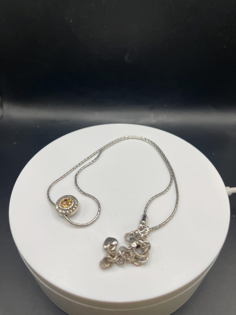 Op de afbeelding: Een zilveren ketting met twee verschillende hangers. E&eacute;n hanger is een cirkelvormig ontwerp met een gele edelsteen in het midden. De andere hanger is een cluster van zilveren vormen. De ketting wordt gepresenteerd op een wit oppervlak tegen een donkere achtergrond.