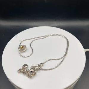 Op de afbeelding: Een zilveren ketting met een kleine, ronde hanger met een gele edelsteen. De ketting is delicaat en de sluiting heeft een decoratief bloemmotief. De ketting wordt gepresenteerd op een wit oppervlak tegen een donkere achtergrond.