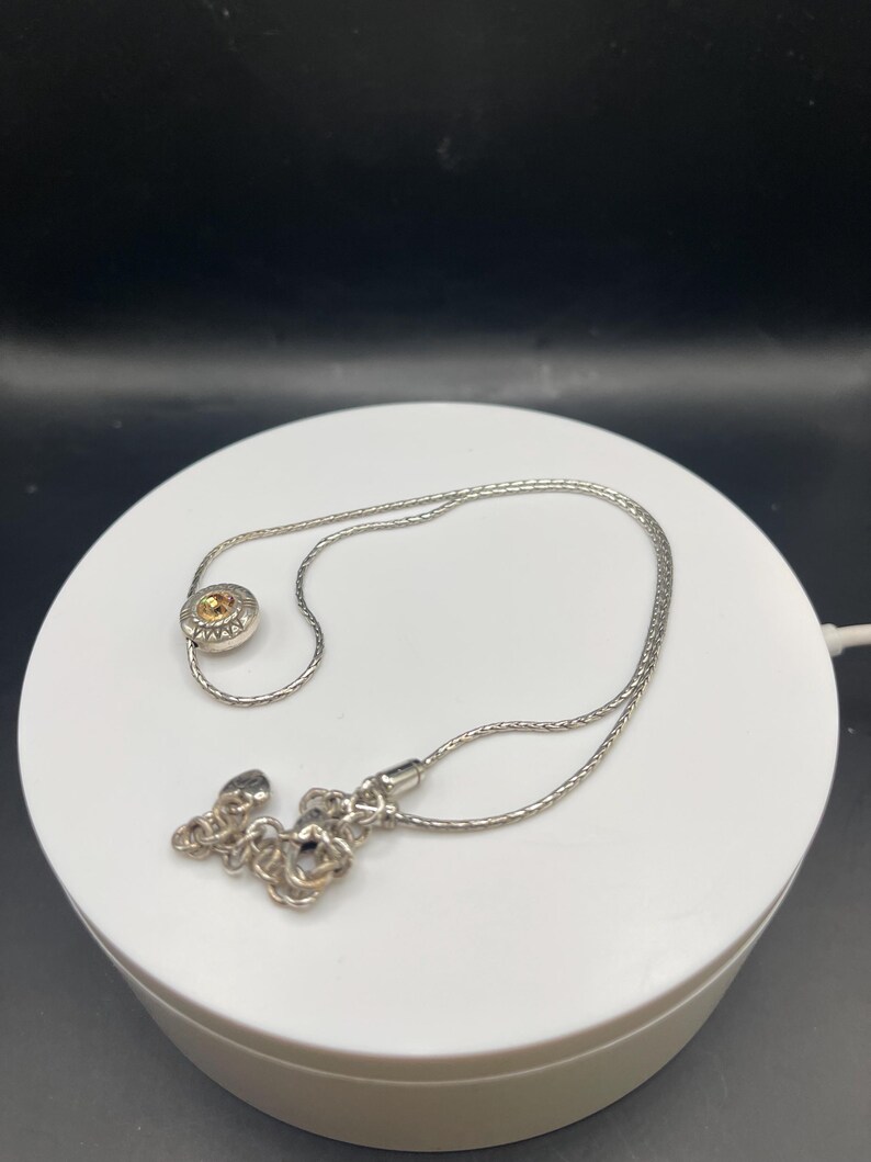 Op de afbeelding: Een zilveren ketting met een kleine, ronde hanger met een gele edelsteen. De ketting is delicaat en de sluiting heeft een decoratief bloemmotief. De ketting wordt gepresenteerd op een wit oppervlak tegen een donkere achtergrond.