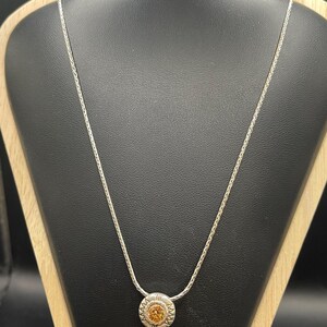 Op de afbeelding: Een zilveren ketting met een ronde hanger met een lichtoranje edelsteen. De hanger is omcirkeld door een decoratief zilveren ontwerp. De ketting wordt gepresenteerd op een zwarte sieradenstandaard.