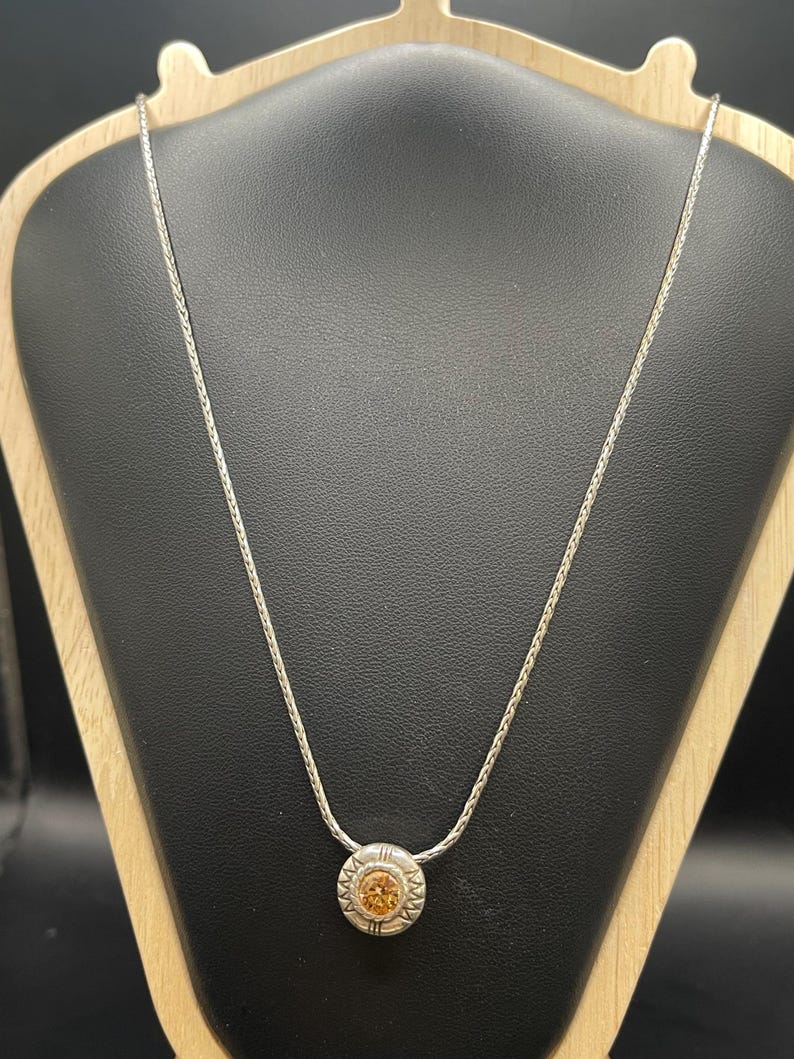 Op de afbeelding: Een zilveren ketting met een ronde hanger met een lichtoranje edelsteen. De hanger is omcirkeld door een decoratief zilveren ontwerp. De ketting wordt gepresenteerd op een zwarte sieradenstandaard.