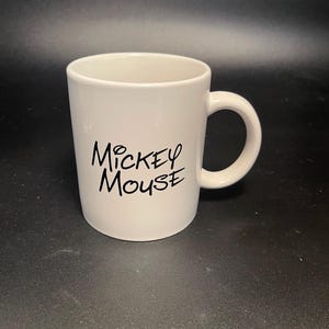 May include: A white ceramic mug with a curved handle. The mug features the words "Mickey Mouse" in black script on the front. The mug is cylindrical and has a smooth, glossy finish.