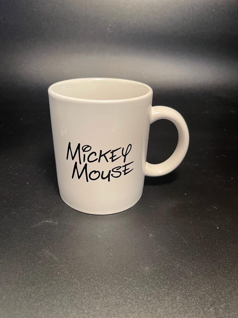 May include: A white ceramic mug with a curved handle. The mug features the words "Mickey Mouse" in black script on the front. The mug is cylindrical and has a smooth, glossy finish.