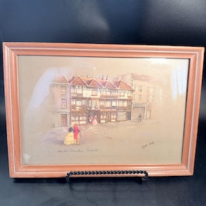 May include: A framed print depicting the Red Lion Hotel in Colchester, featuring a timber-framed building with figures in period clothing in the foreground. The artwork is signed by the artist and includes the text The Red Lion Inn, Colchester.