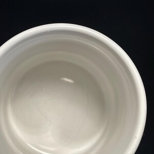 May include: A white ceramic bowl with a rounded rim. The interior of the bowl is smooth and reflective, with a slight curve. The bowl is set against a black background.