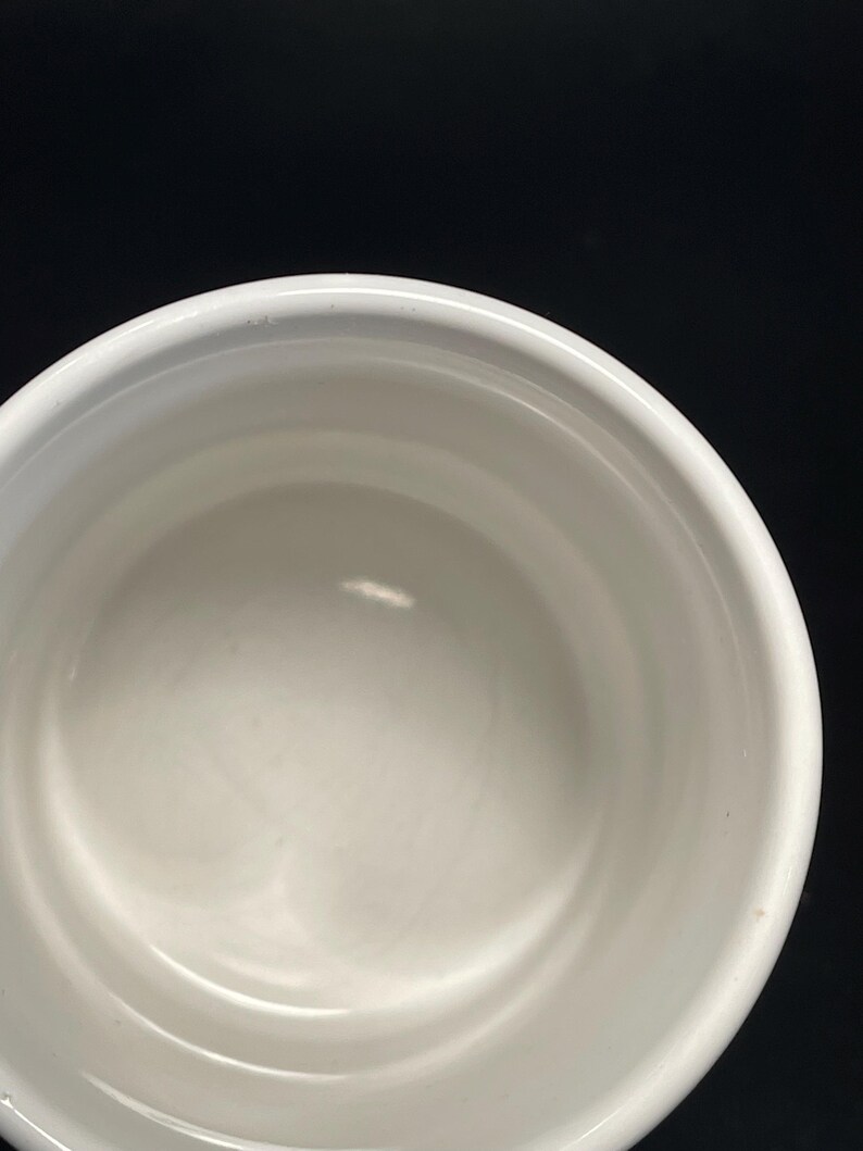 May include: A white ceramic bowl with a rounded rim. The interior of the bowl is smooth and reflective, with a slight curve. The bowl is set against a black background.