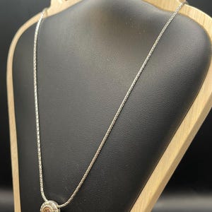 Op de afbeelding: Zilverkleurige ketting met een ronde hanger. De hanger heeft een centrale, lichtgekleurde steen, omringd door een gedetailleerd, zon-achtig ontwerp. De ketting wordt getoond op een zwarte sieradenstandaard met een houten frame.