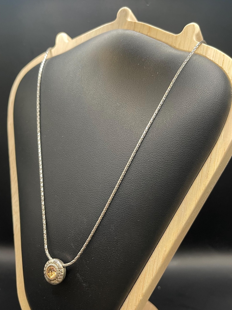 Op de afbeelding: Zilverkleurige ketting met een ronde hanger. De hanger heeft een centrale, lichtgekleurde steen, omringd door een gedetailleerd, zon-achtig ontwerp. De ketting wordt getoond op een zwarte sieradenstandaard met een houten frame.