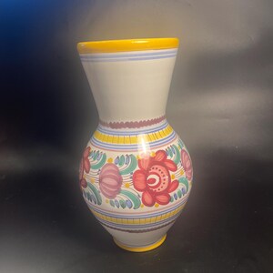 May include: A ceramic vase with a white base, featuring a yellow rim and base. The body is decorated with horizontal bands of blue and brown lines, interspersed with hand-painted floral motifs in red, pink, and green, creating a symmetrical pattern.